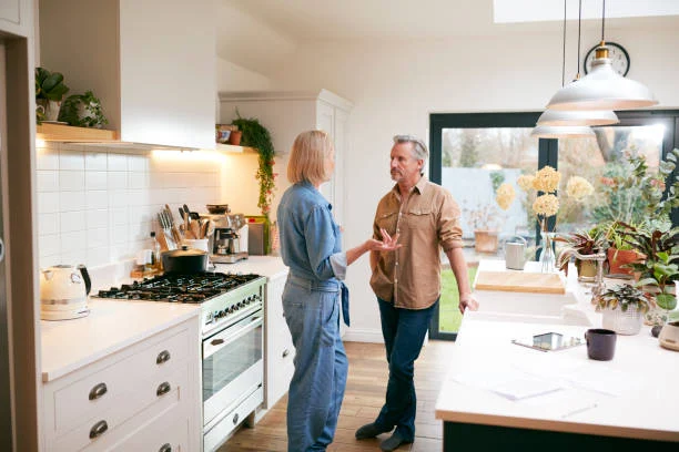 Two adults talking in a bright kitchen with a white stove, pendant lights and potted plants beside a large window