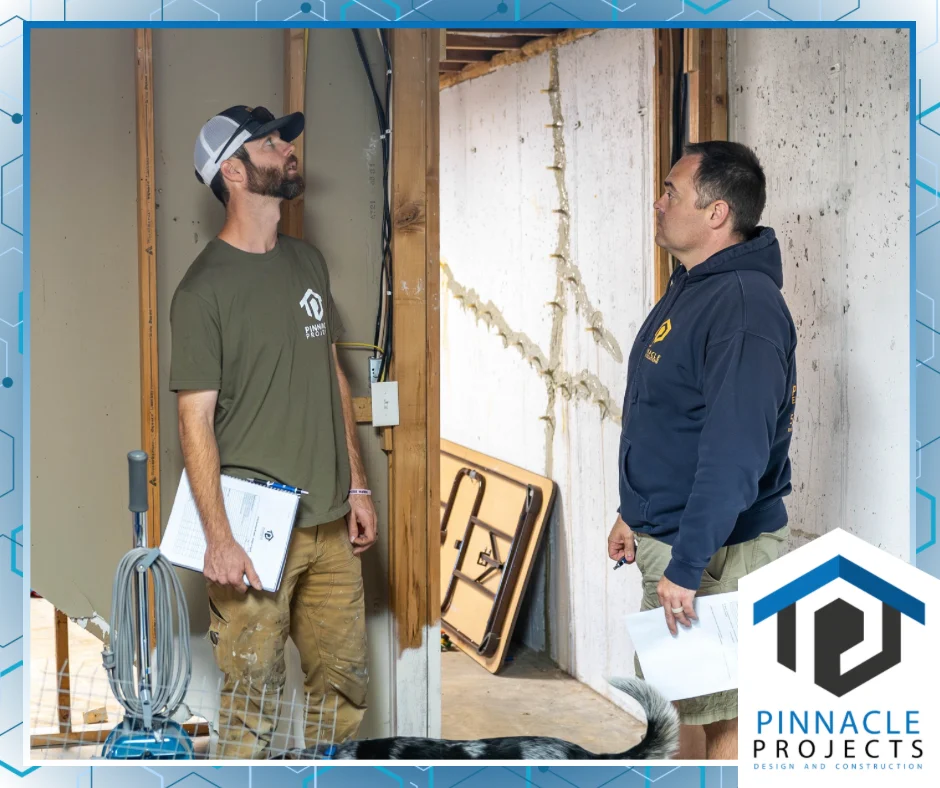 Two men with paperwork standing among exposed studs and concrete walls, with the Pinnacle Projects Design and Construction logo