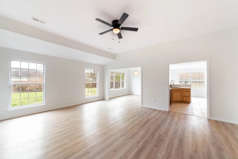 Empty room with wood-look flooring, white walls, large windows, a ceiling fan and doorways to a kitchen and dining area