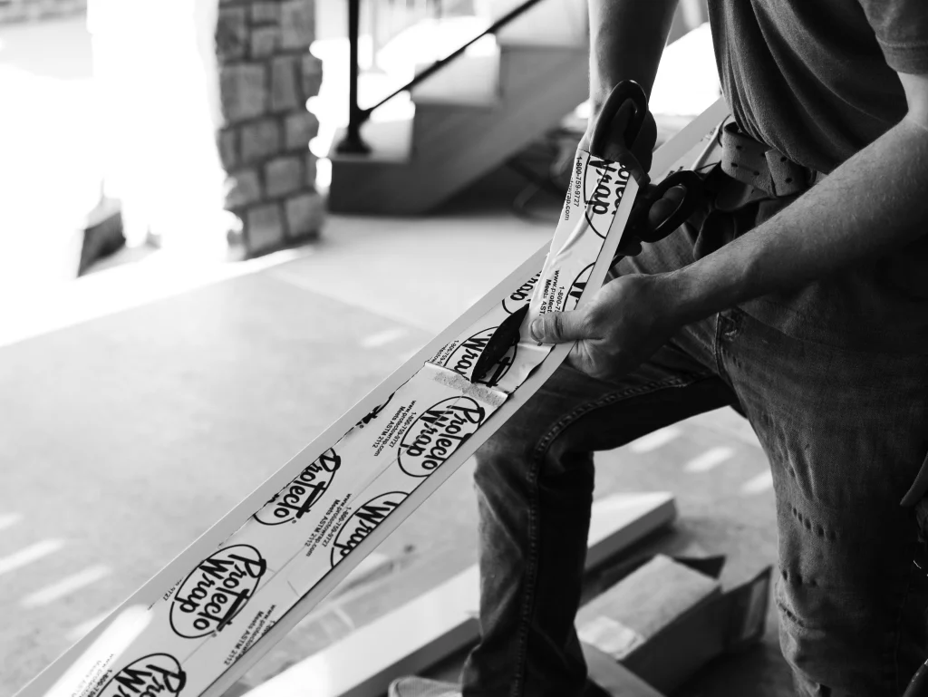 Black-and-white photo of a person using large scissors to cut material labeled Protecto Wrap along a long frame
