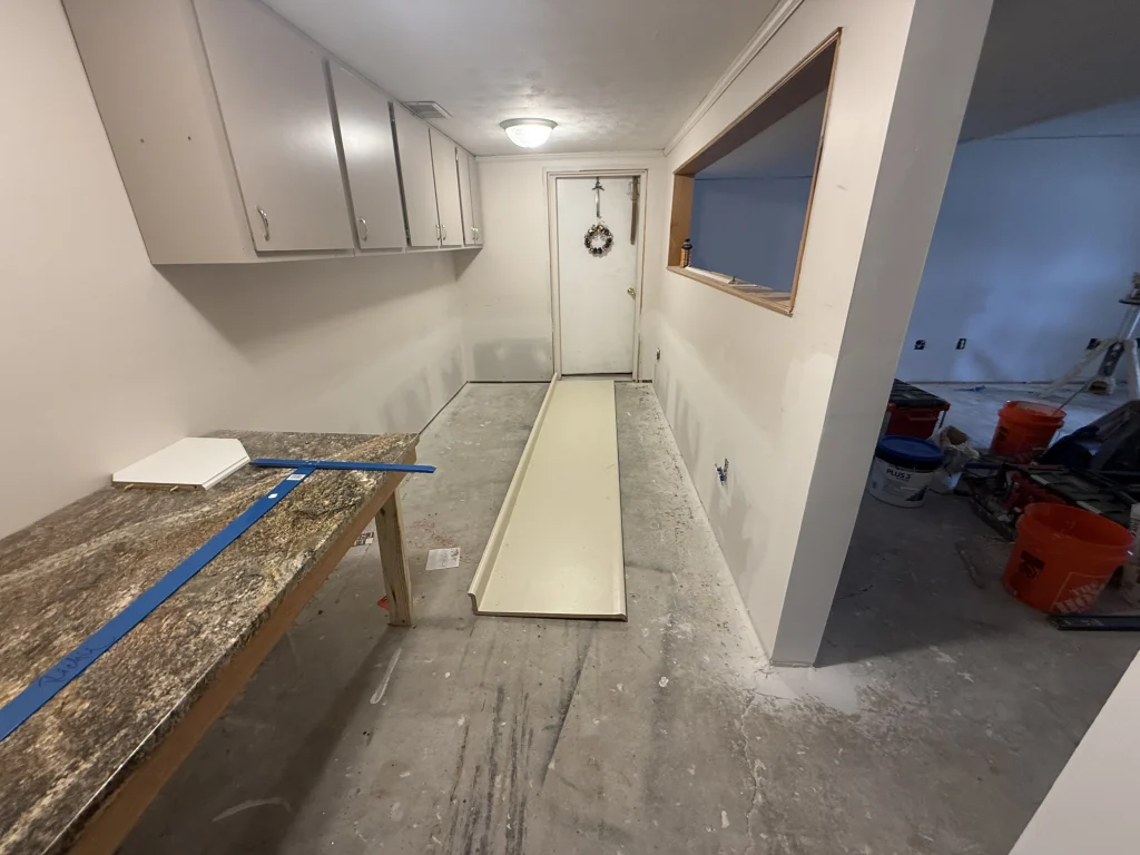 Room under renovation with wall cabinets, a worktop, a loose panel on the bare floor and a bucket labeled Plus 3