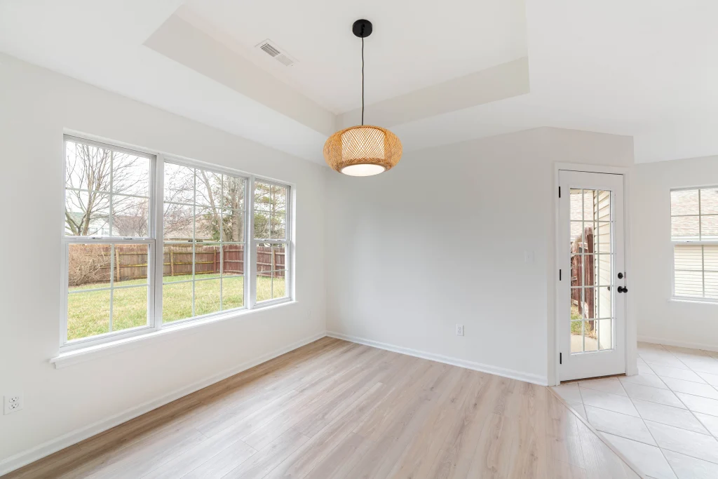 Empty white room with wood flooring, large windows, a woven pendant light and a glass-paneled door beside tile flooring