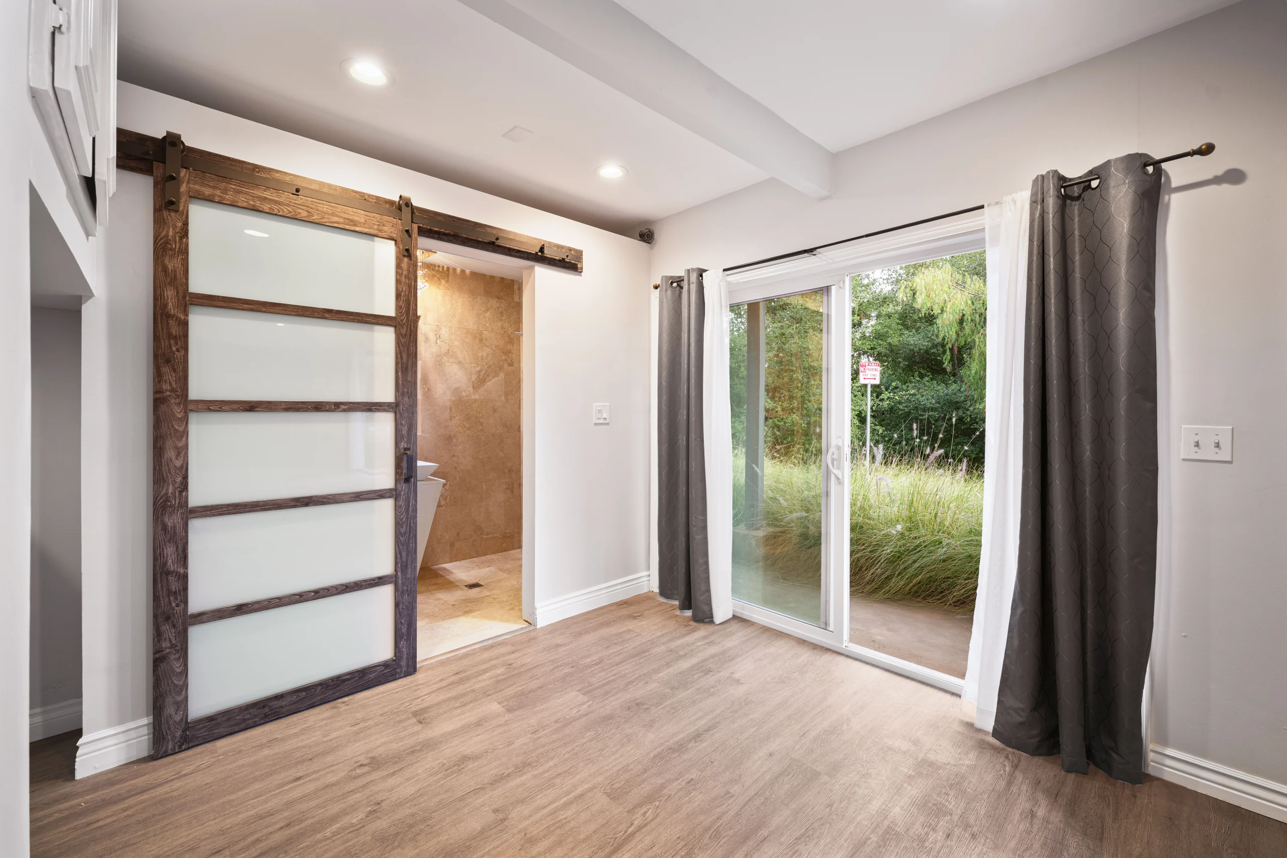 Empty room with wood flooring, rustic sliding barn door, recessed lights, and glass patio doors with gray curtains