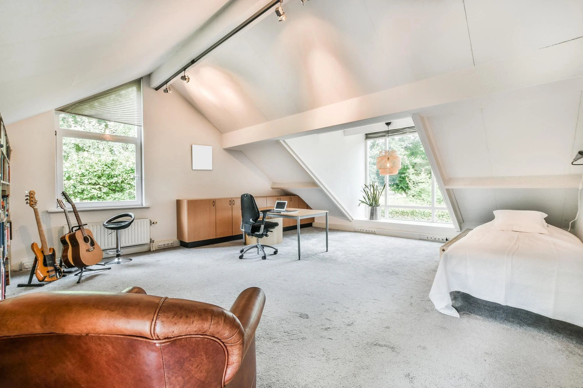 Bright attic room with sloped ceilings, large windows, a bed, desk, office chair, guitars, and brown leather seating
