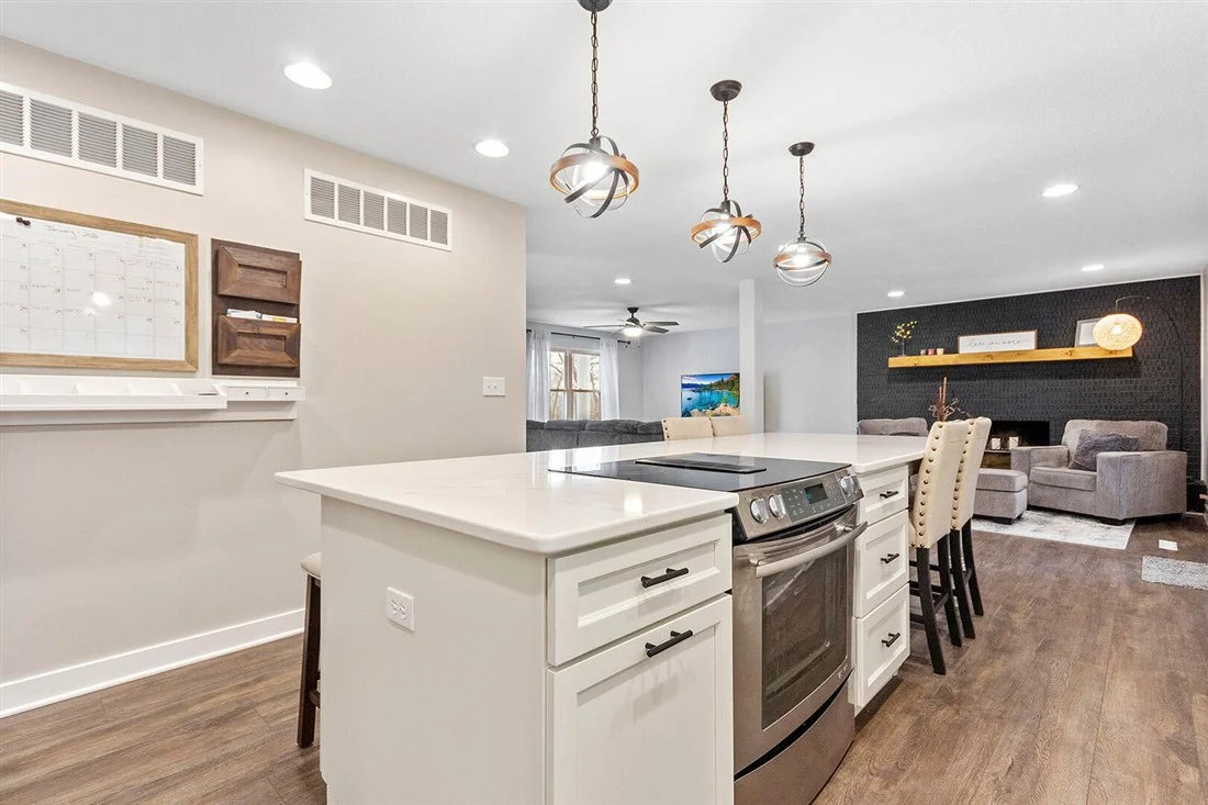 White kitchen island with stainless steel stove, pendant lights and stools; gray armchairs beyond