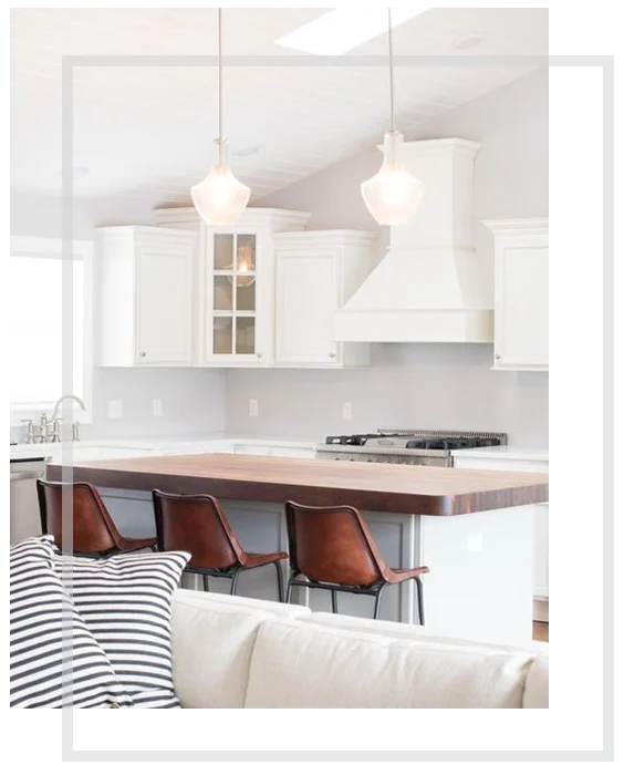 White kitchen with pendant lights, wood-topped island and three brown stools; sofa and striped cushions in foreground