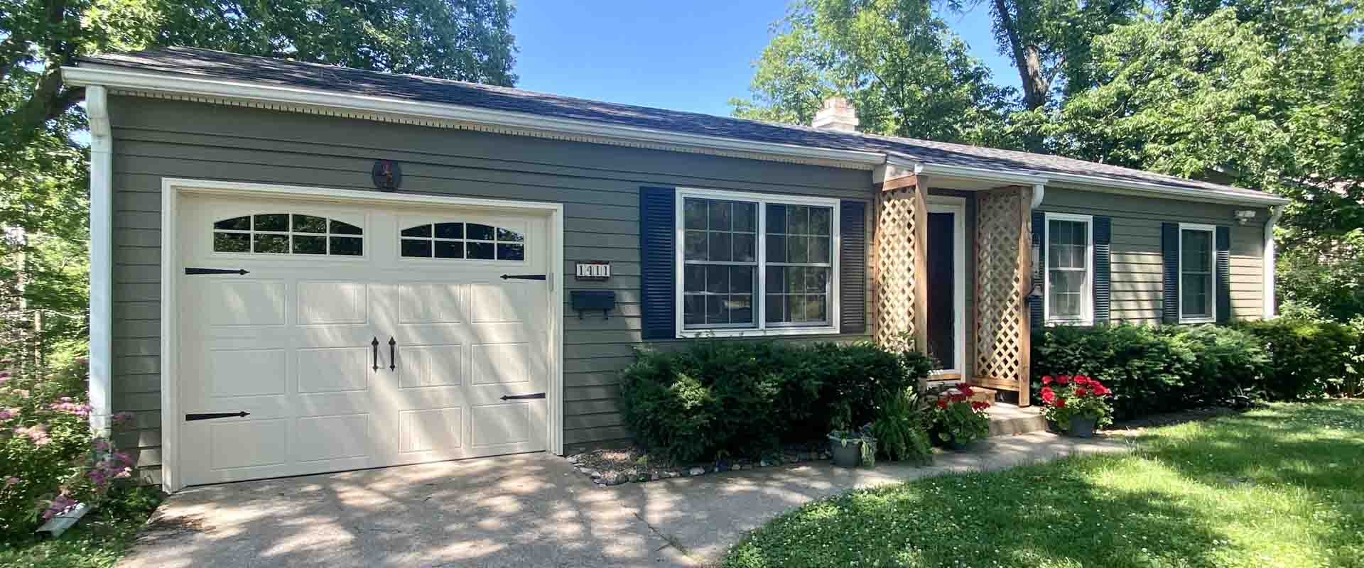 Single-story gray house with an attached garage, dark shutters, lattice porch posts, shrubs and a lawn