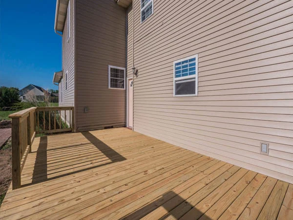 Empty wooden deck with railing beside a beige-sided house with windows, under a clear blue sky