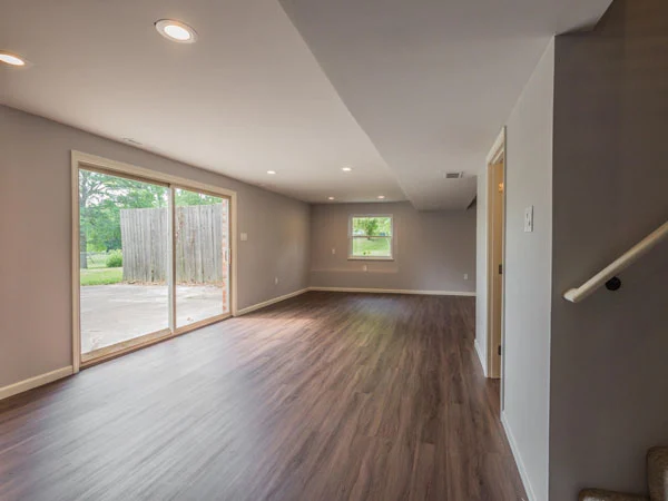 Empty gray room with wood-look flooring, sliding glass patio doors, a small window, and stairs at right