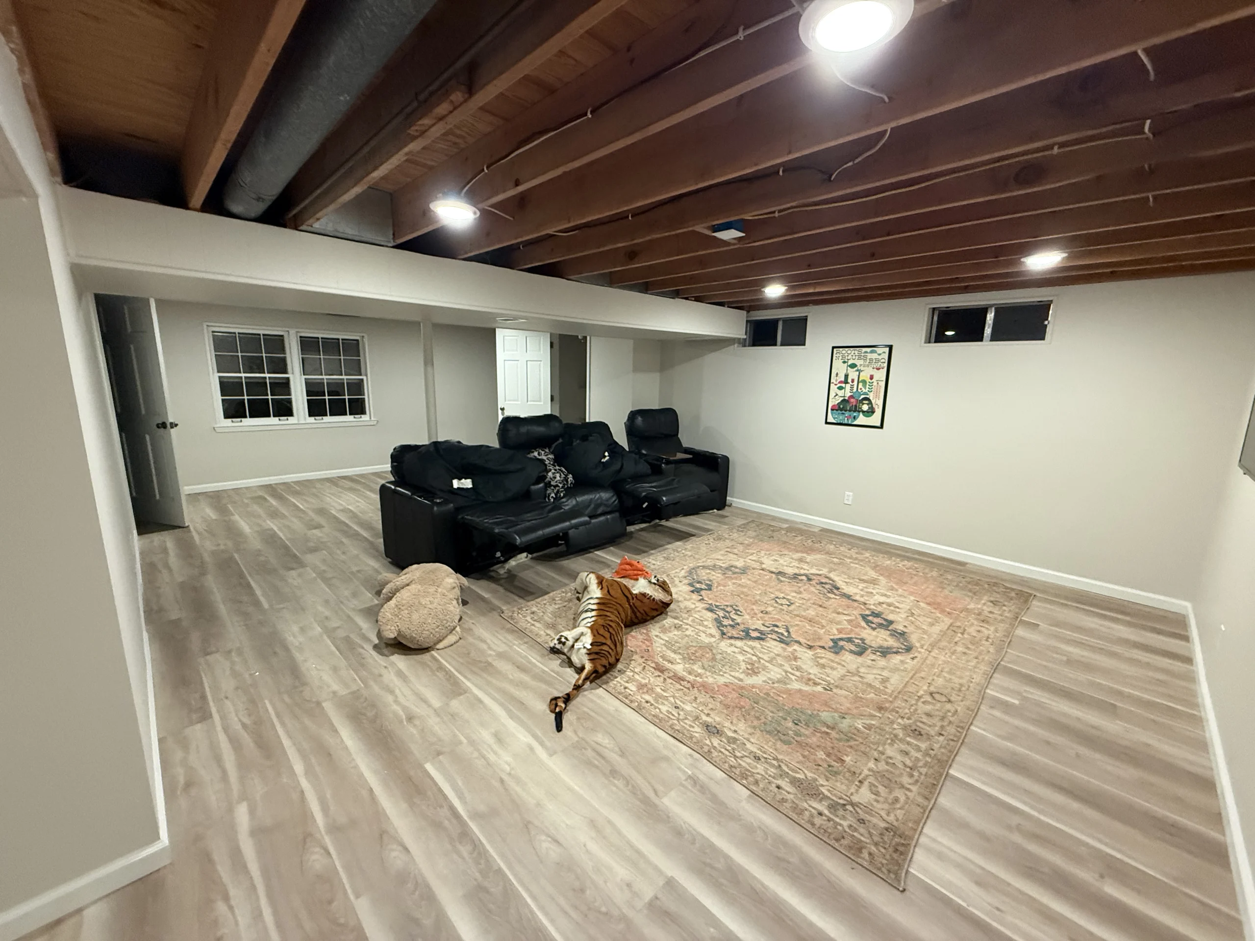 Room with exposed ceiling joists, black reclining seats, patterned rug, stuffed tiger, windows and framed wall art