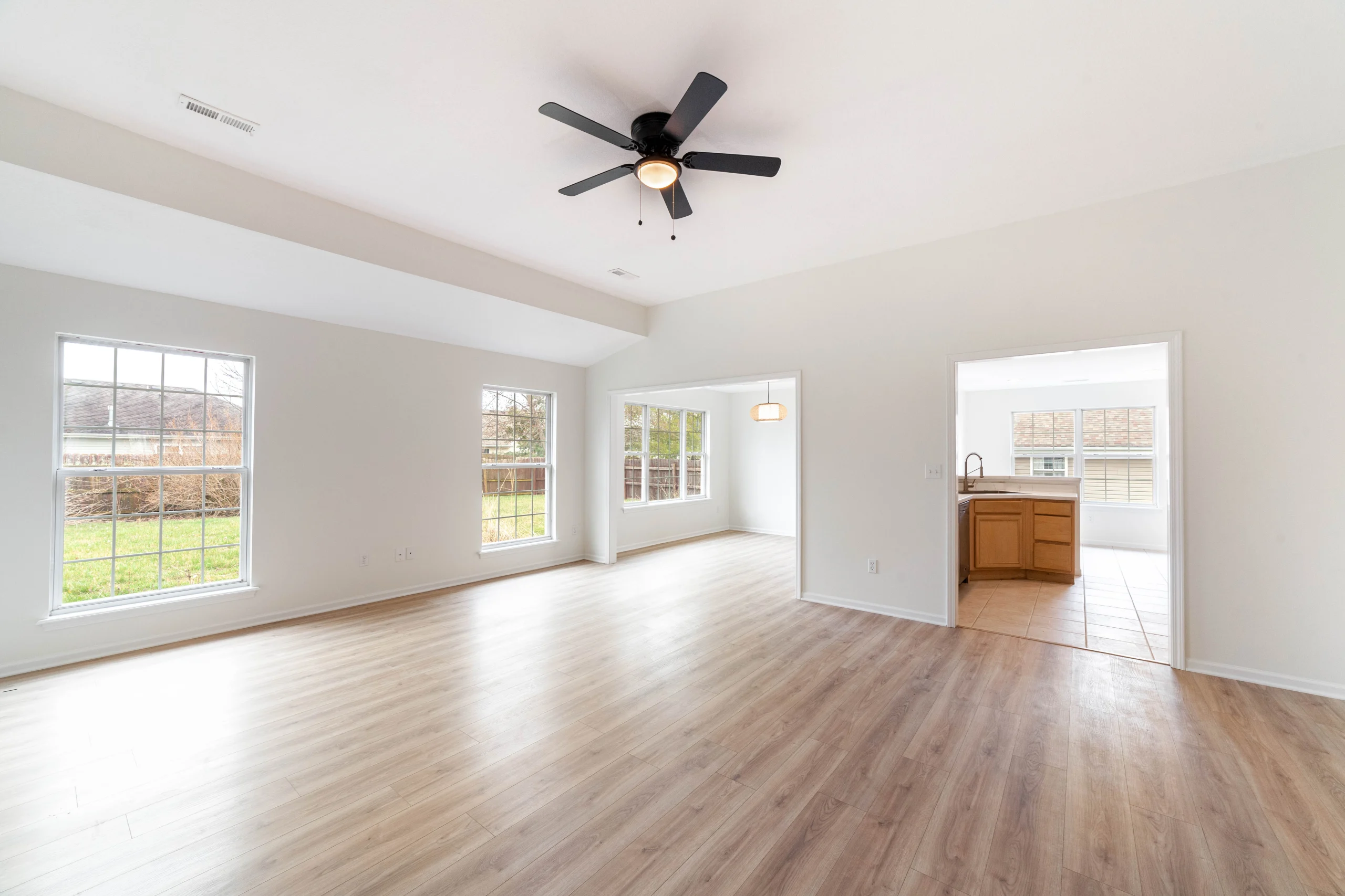 Empty room with wood-look flooring, large windows, a black ceiling fan and doorways to adjoining rooms and a kitchen