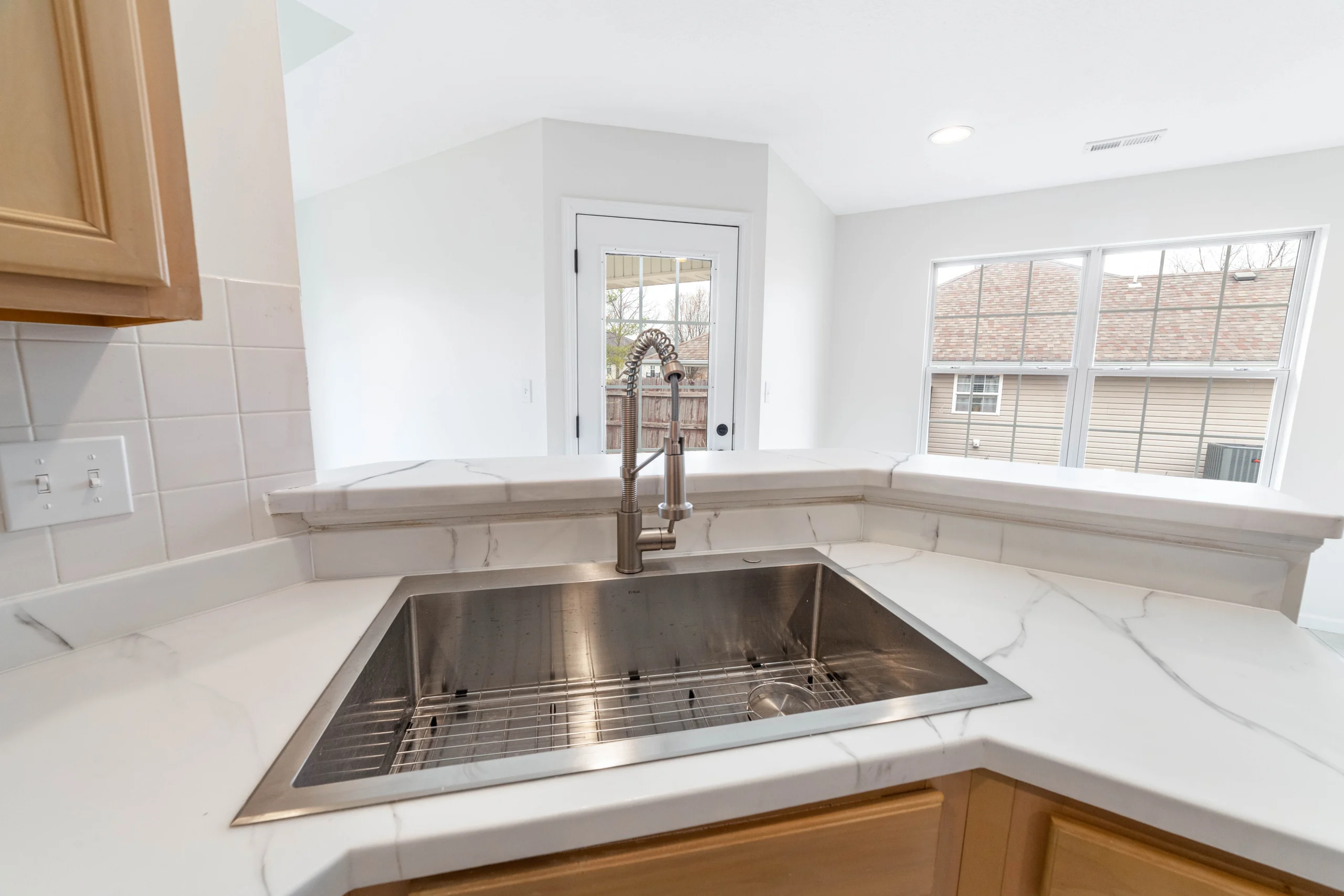 Stainless steel sink with tall spring faucet, white veined counters, wood cabinets, windows and a glass-paneled door
