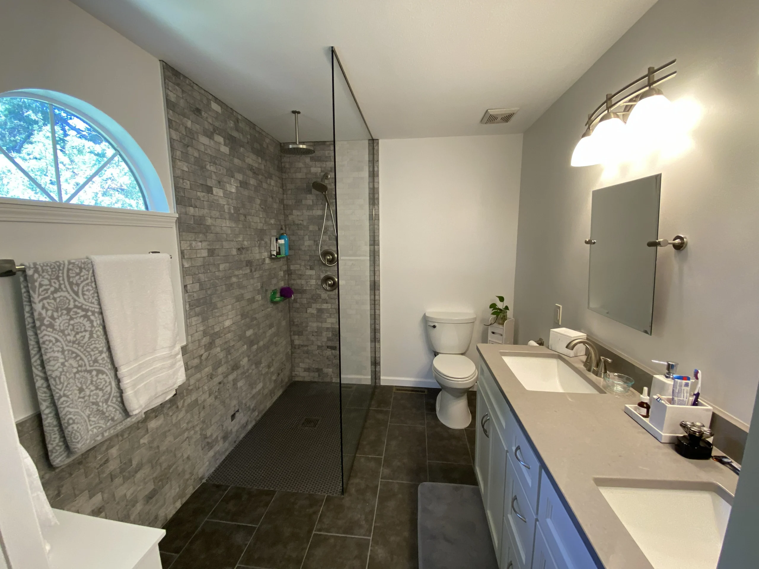 Bathroom with glass shower partition, gray tiled walls, toilet, double-sink vanity, mirror, towels, and arched window