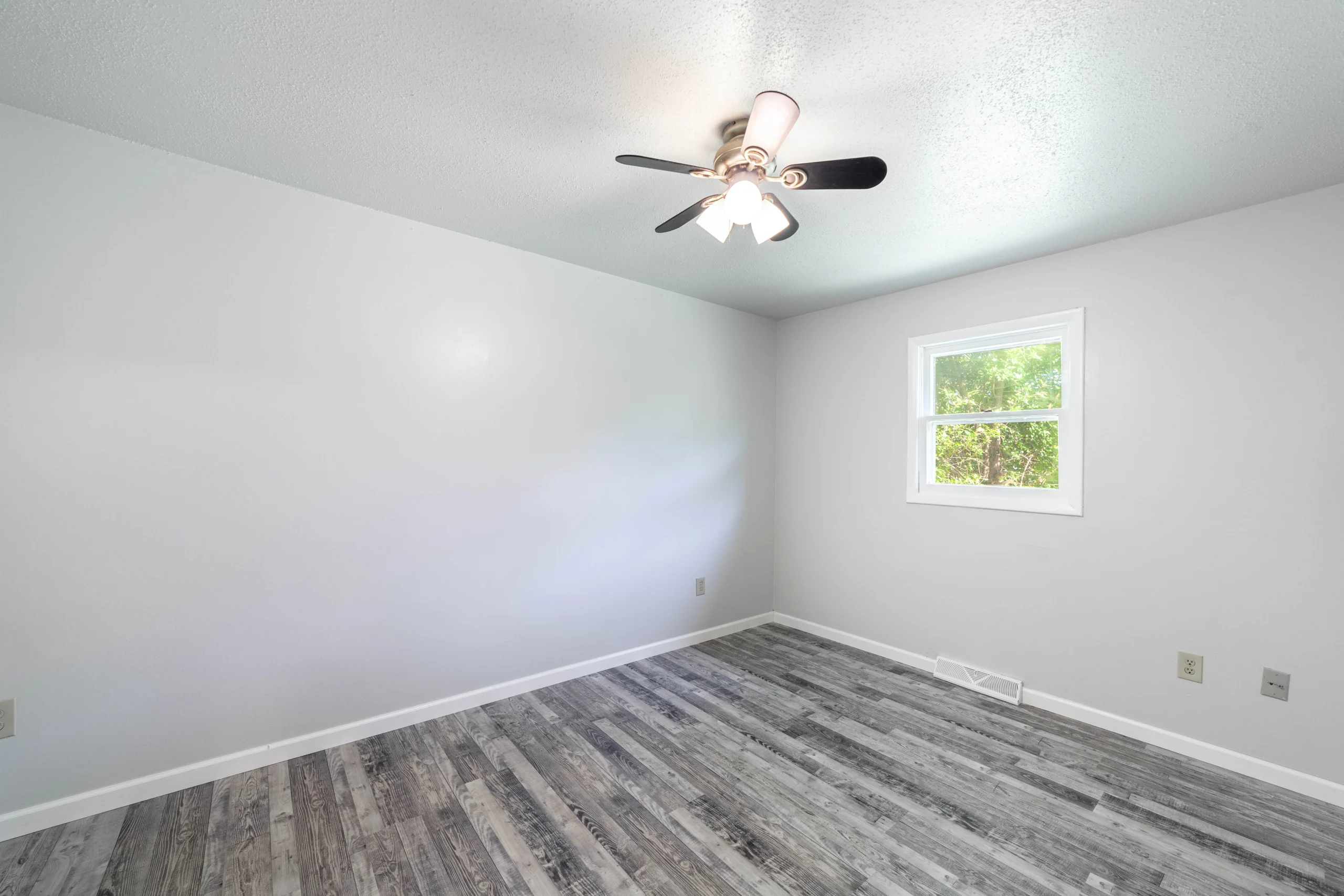 Empty room with light gray walls, gray wood-look flooring, one window, and a ceiling fan with lights on
