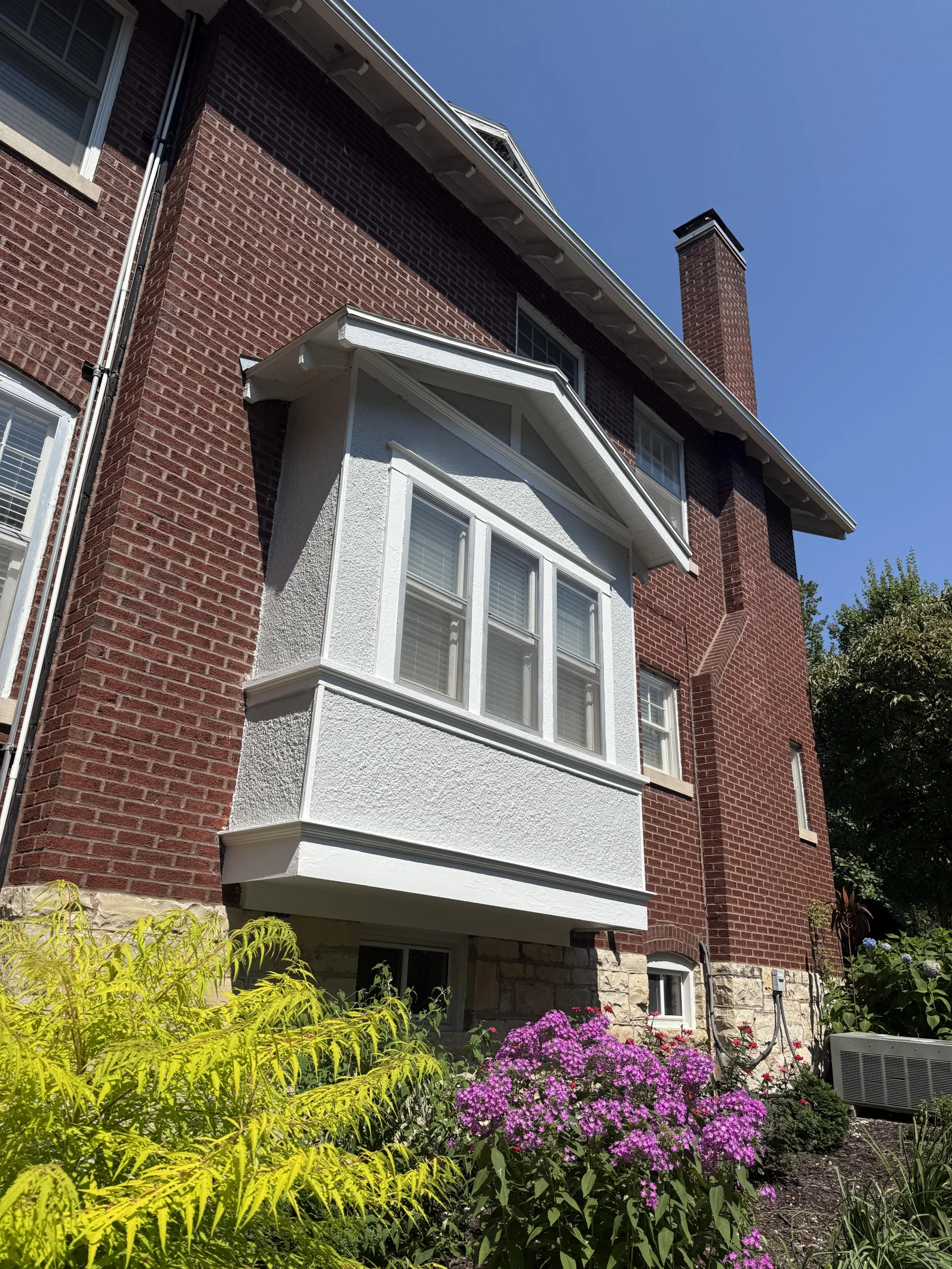 Red-brick house with a projecting gray bay window, chimney, and garden with yellow-green foliage and purple flowers