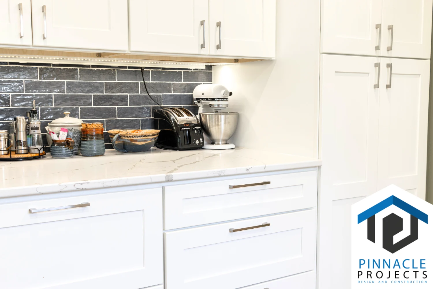 White kitchen cabinets, dark tile backsplash, toaster and mixer; logo “PINNACLE PROJECTS DESIGN AND CONSTRUCTION”