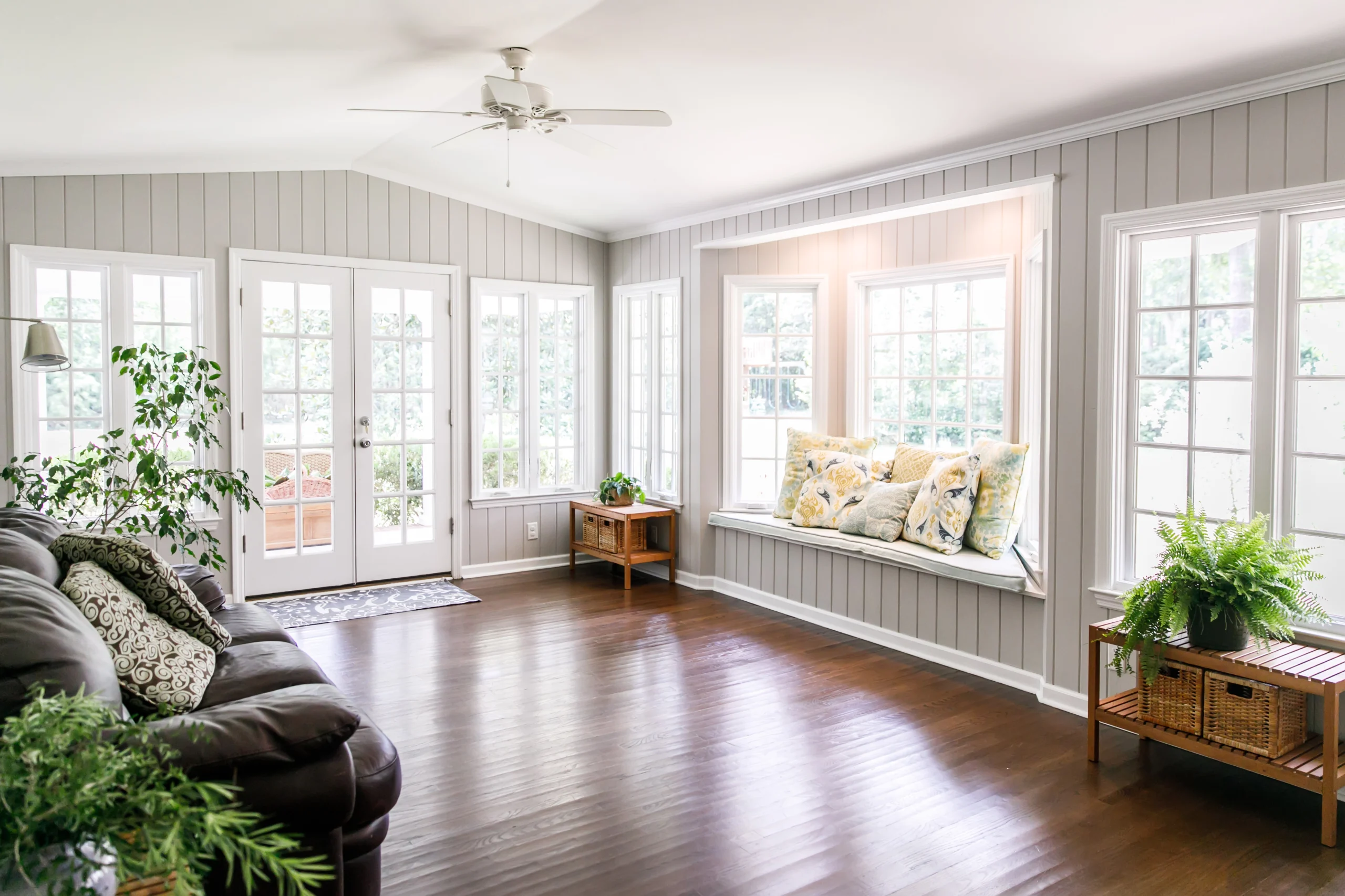 Sunlit room with many windows, gray paneled walls, wood floor, dark sofa, cushioned window seat, plants and ceiling fan