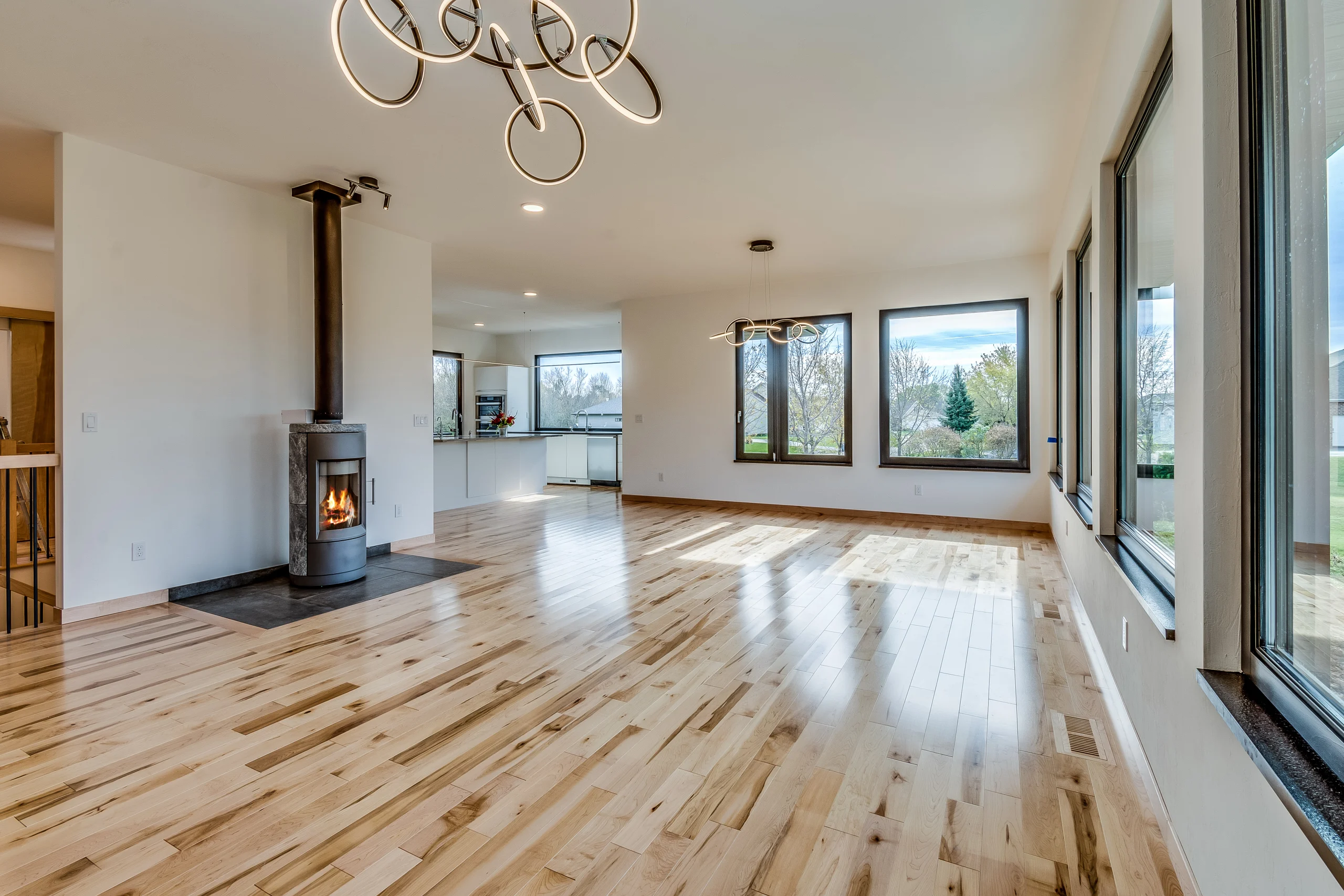 Empty room with light wood flooring, a lit wood stove, looping ceiling lights, large windows and an adjoining kitchen