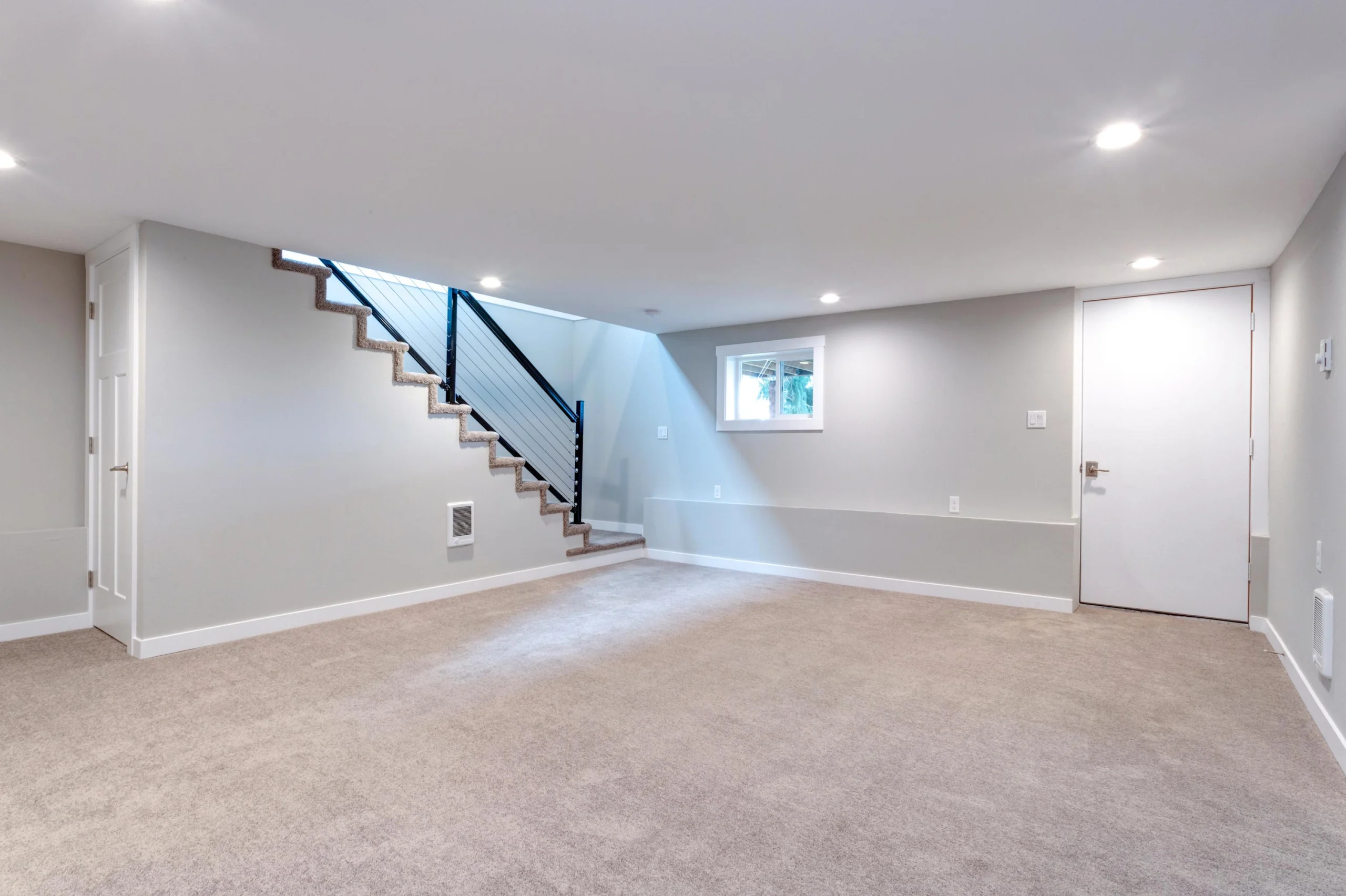 Empty gray room with beige carpet, stairs with black railing, a small window, recessed lights, and white doors