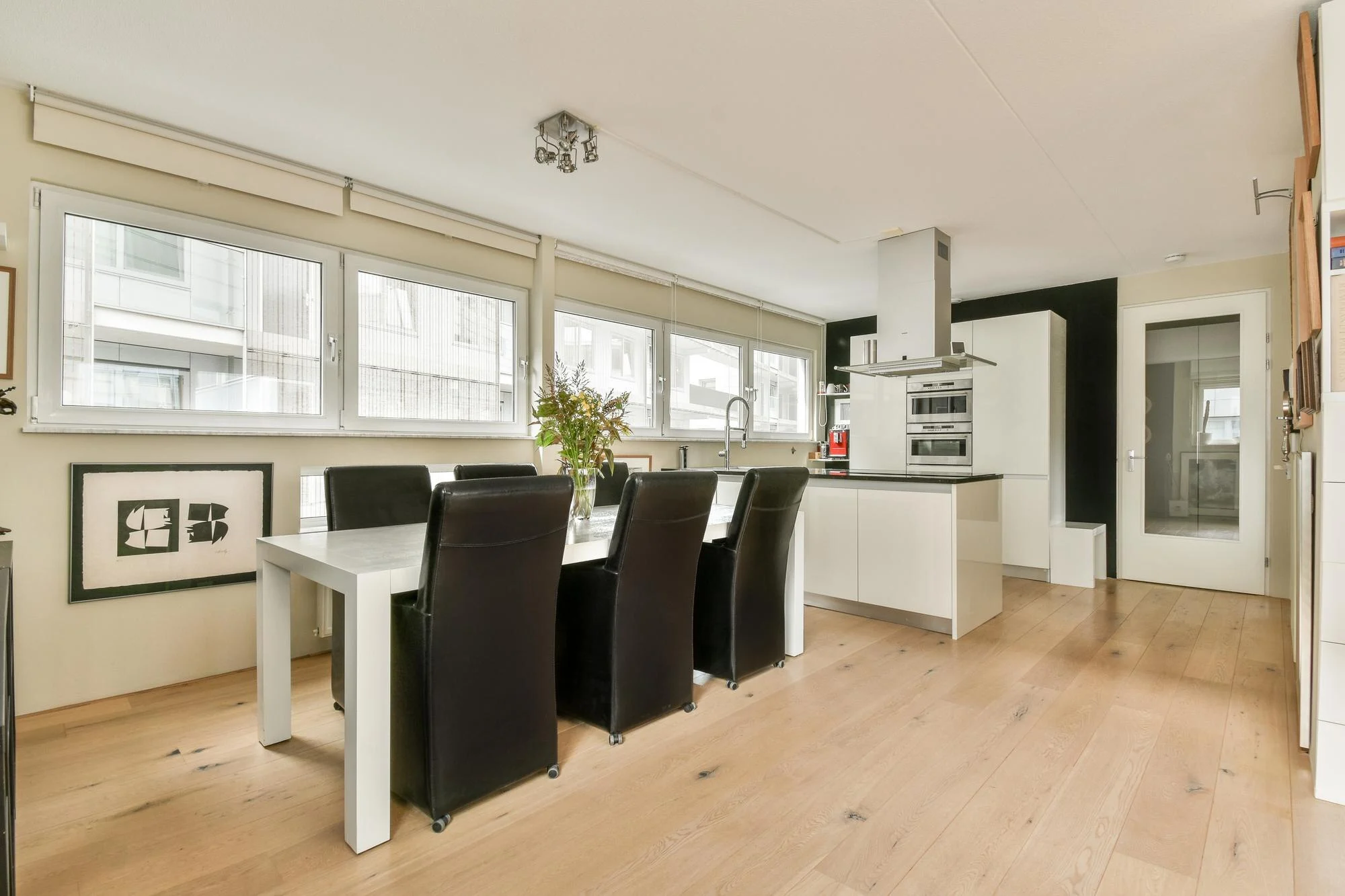 Bright kitchen and dining area with white table, black chairs, large windows, white cabinets, and wood flooring