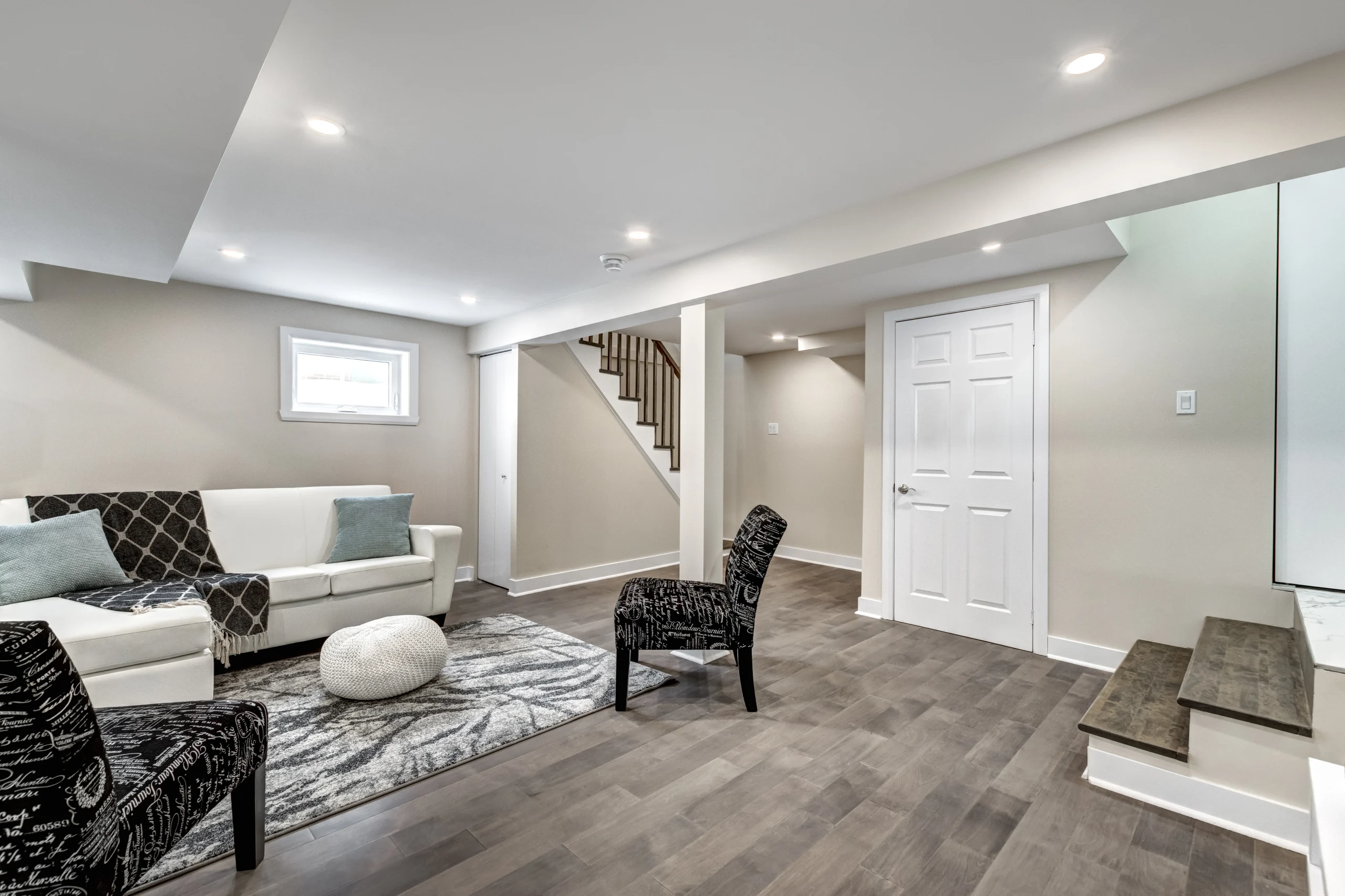 Room with white sofa, patterned chairs, rugs, round pouf, gray flooring, stairs, white doors and recessed lights