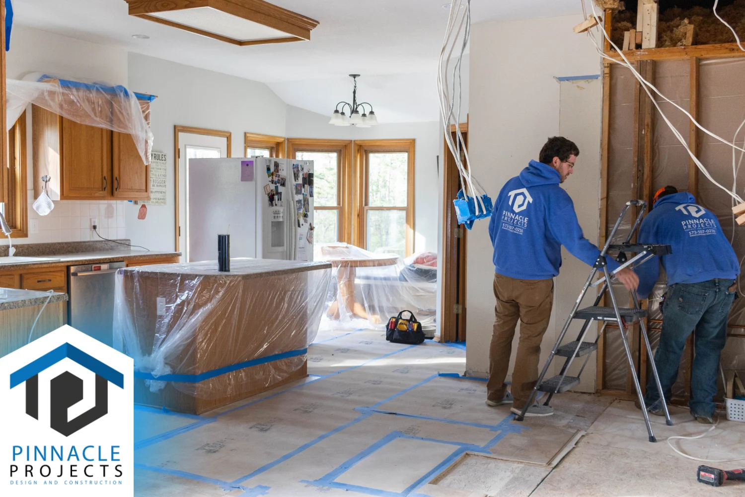 Two workers in blue hoodies beside a ladder in a kitchen with exposed framing; logo reads “PINNACLE PROJECTS”