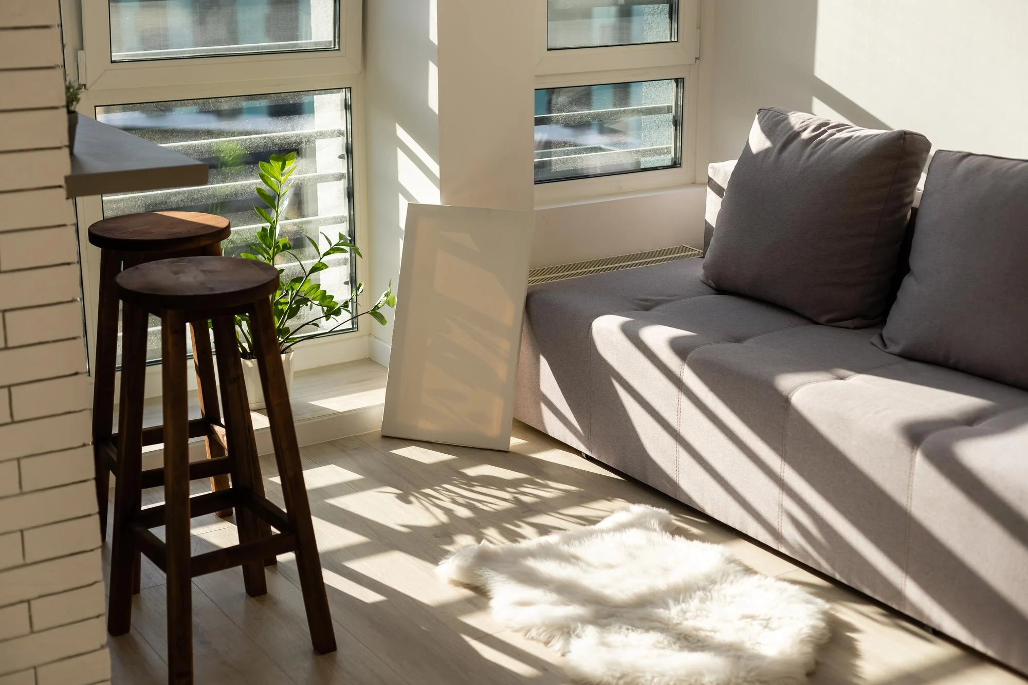 Sunlit room with a gray sofa, two wooden stools, a plant, a leaning frame, large windows and a white rug