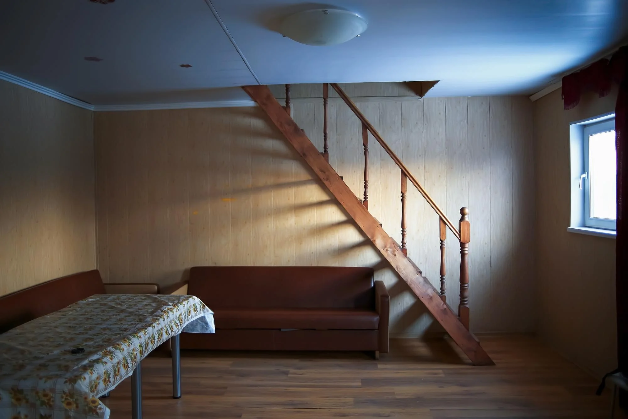 Dim room with a wooden staircase, brown sofa, cloth-covered table, window and wood-look floor